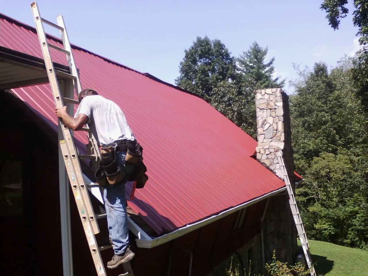 Roofer on a ladder beside a red metal roof and stone chimney — Soffit & Fascia Repair by Roofing Brothers in Argos